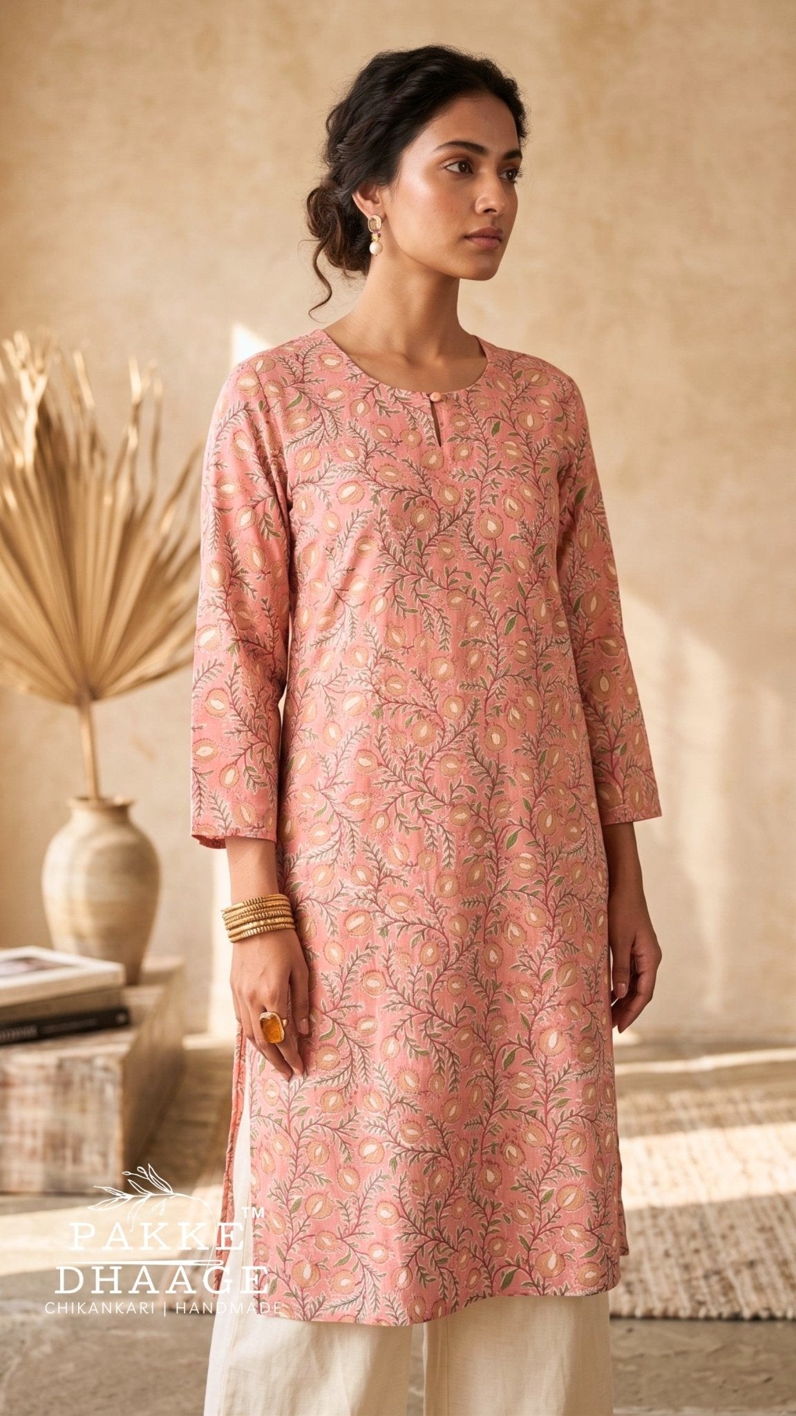 A woman wearing a dusty pink printed kurta with three-quarter sleeves and cream pants, standing in a warm, rustic indoor setting with a textured beige wall and dried palm leaves. | Pakke Dhaage