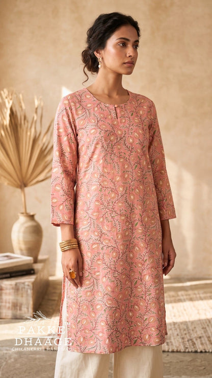 A woman wearing a dusty pink printed kurta with three-quarter sleeves and cream pants, standing in a warm, rustic indoor setting with a textured beige wall and dried palm leaves. | Pakke Dhaage