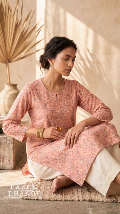 A woman sits cross-legged on a woven mat in a warm, sunlit room wearing a dusty pink printed kurta with white pants. The brand name 'PAKKE DHAAGE' is visible in the bottom-left corner.