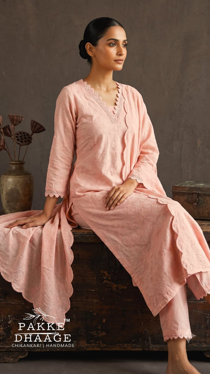 A woman wearing a light pink three-piece salwar kameez set sits on a rustic wooden chest against a textured gray wall. The set includes a long-sleeve tunic with scalloped edges, matching trousers, and a draped dupatta.