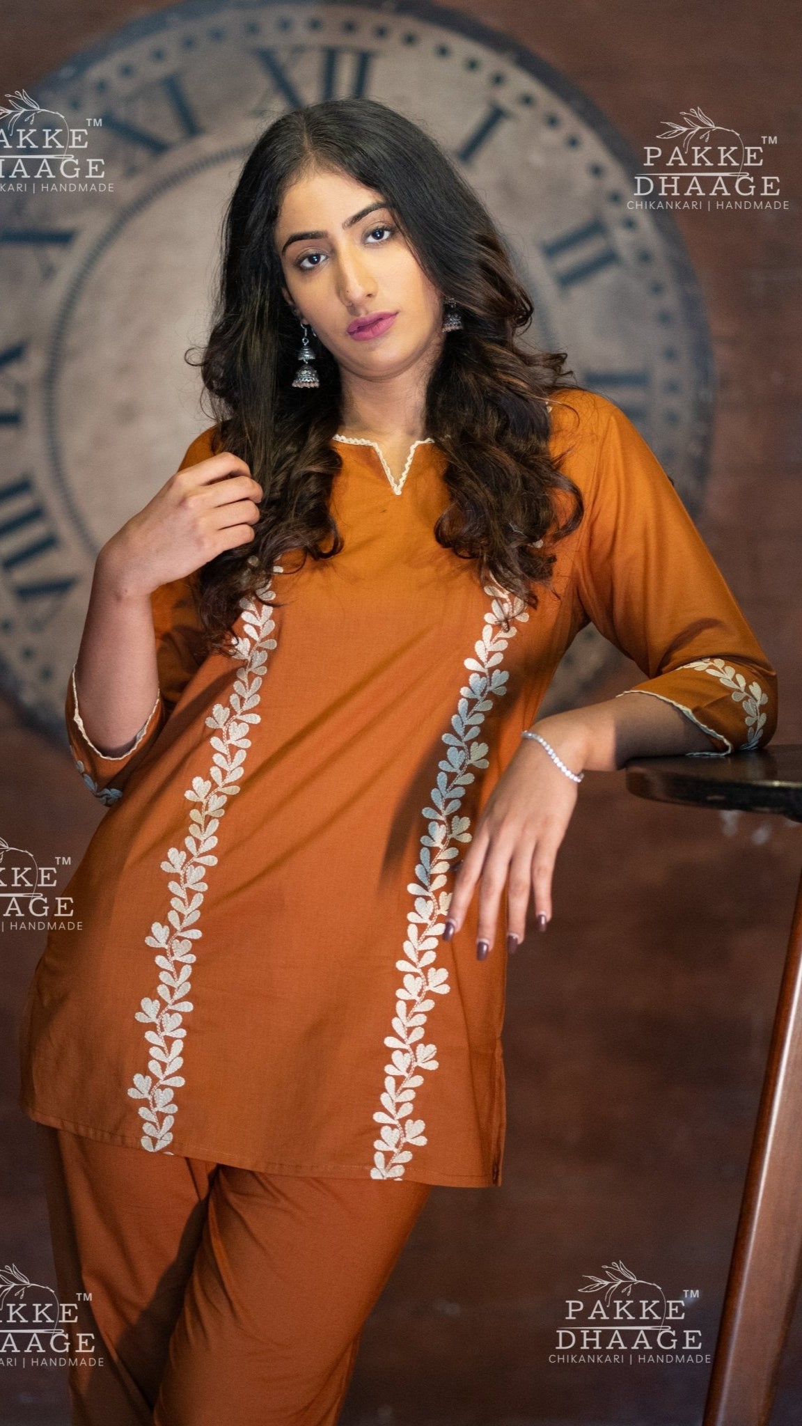 Woman in an orange co-ord set with white embroidery against a decorative wall.