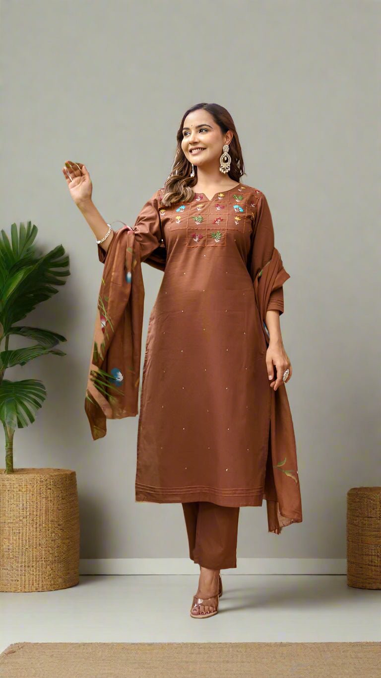 Woman in a brown traditional kurti set with matching dupatta standing in an outdoor setting with plants and a wooden door.
