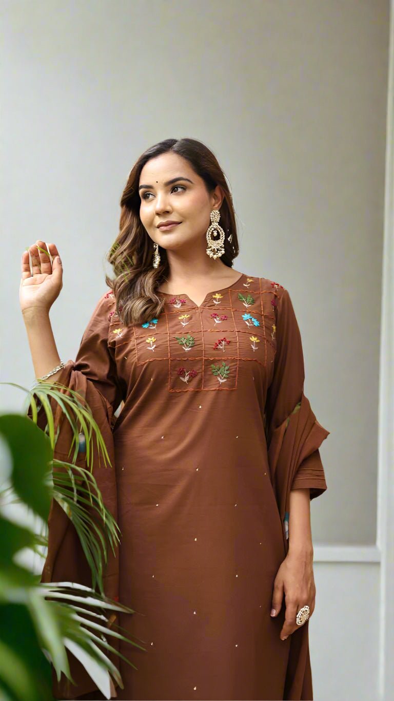 Woman in a brown dress with floral embroidery standing in a garden setting.