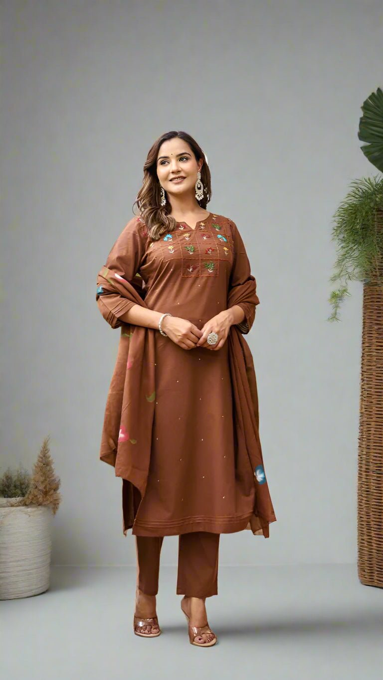 Woman in a brown traditional kurta set standing in an outdoor setting with plants and a wooden door.