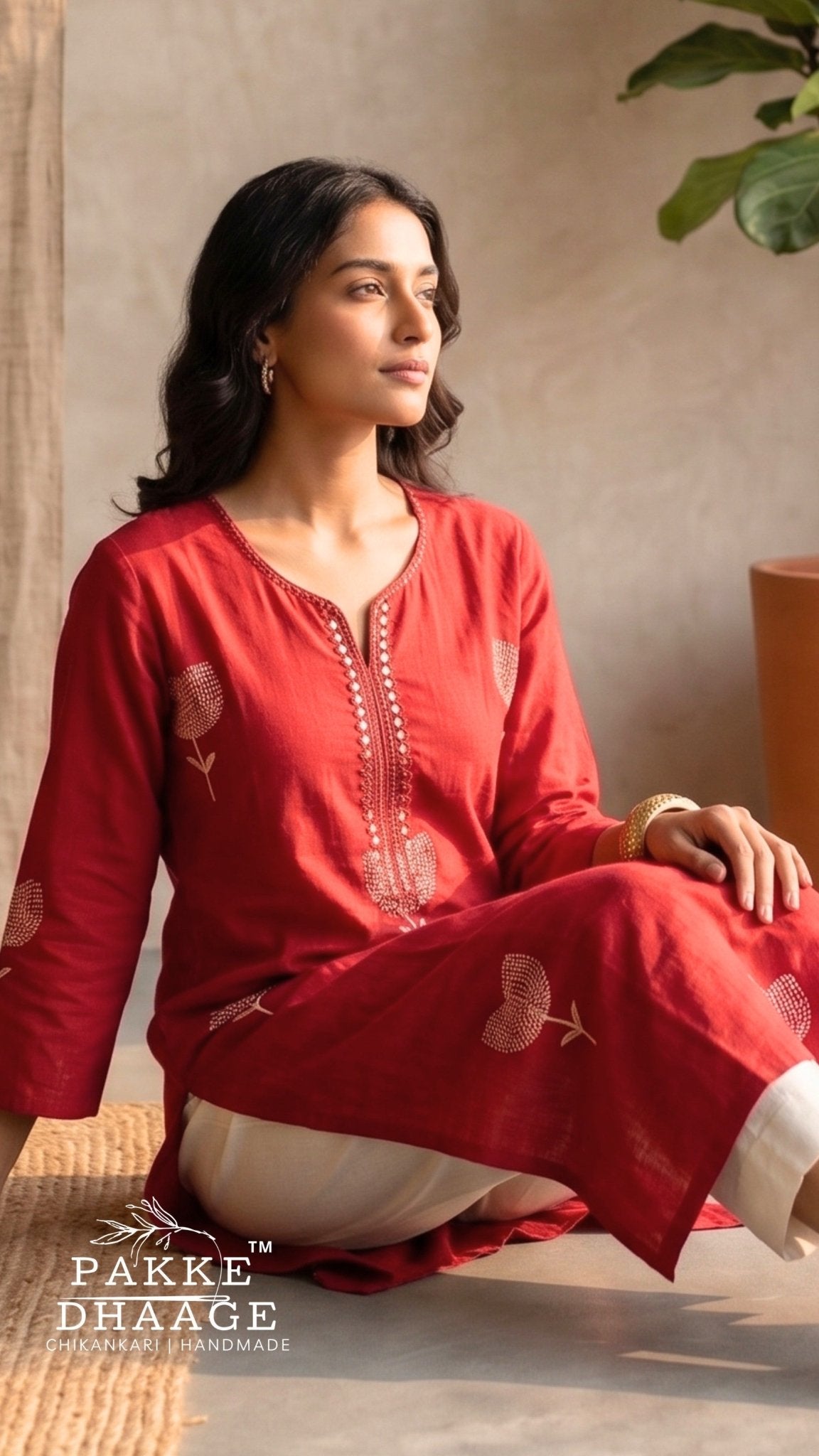 A woman wearing a red long-sleeve kurta with white pants sits on a woven mat in a warm indoor setting. The kurta features embroidered detailing on the chest and sleeves. | Pakke Dhaage