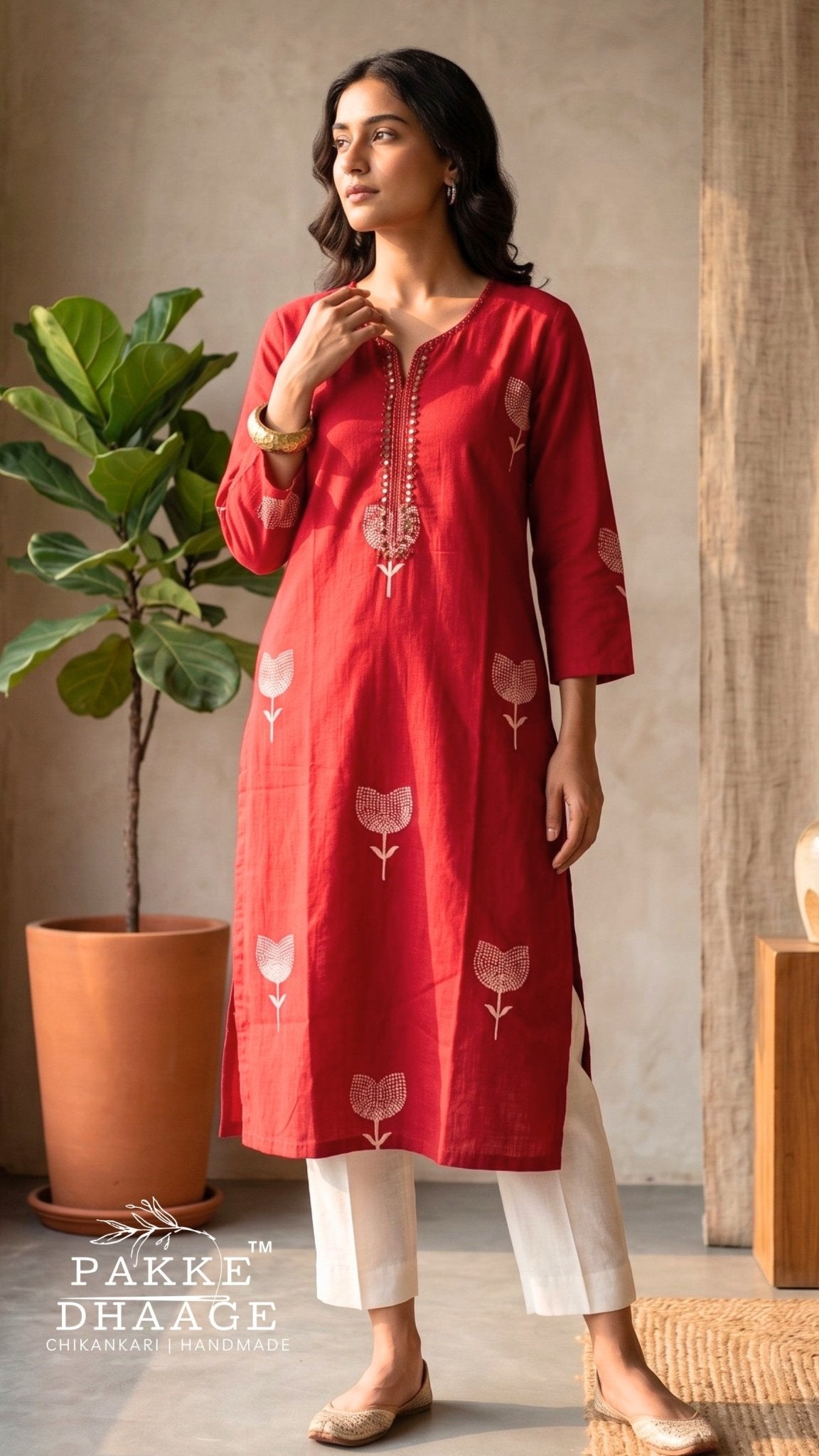 A woman modeling a red three-quarter sleeve kurta with spaced white floral prints and white pants in a styled indoor setting with a potted plant. | Pakke Dhaage