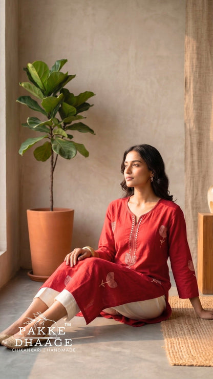 A woman sits on a woven mat in a warm, minimalist room wearing a red embroidered kurta and cream trousers. | Pakke Dhaage