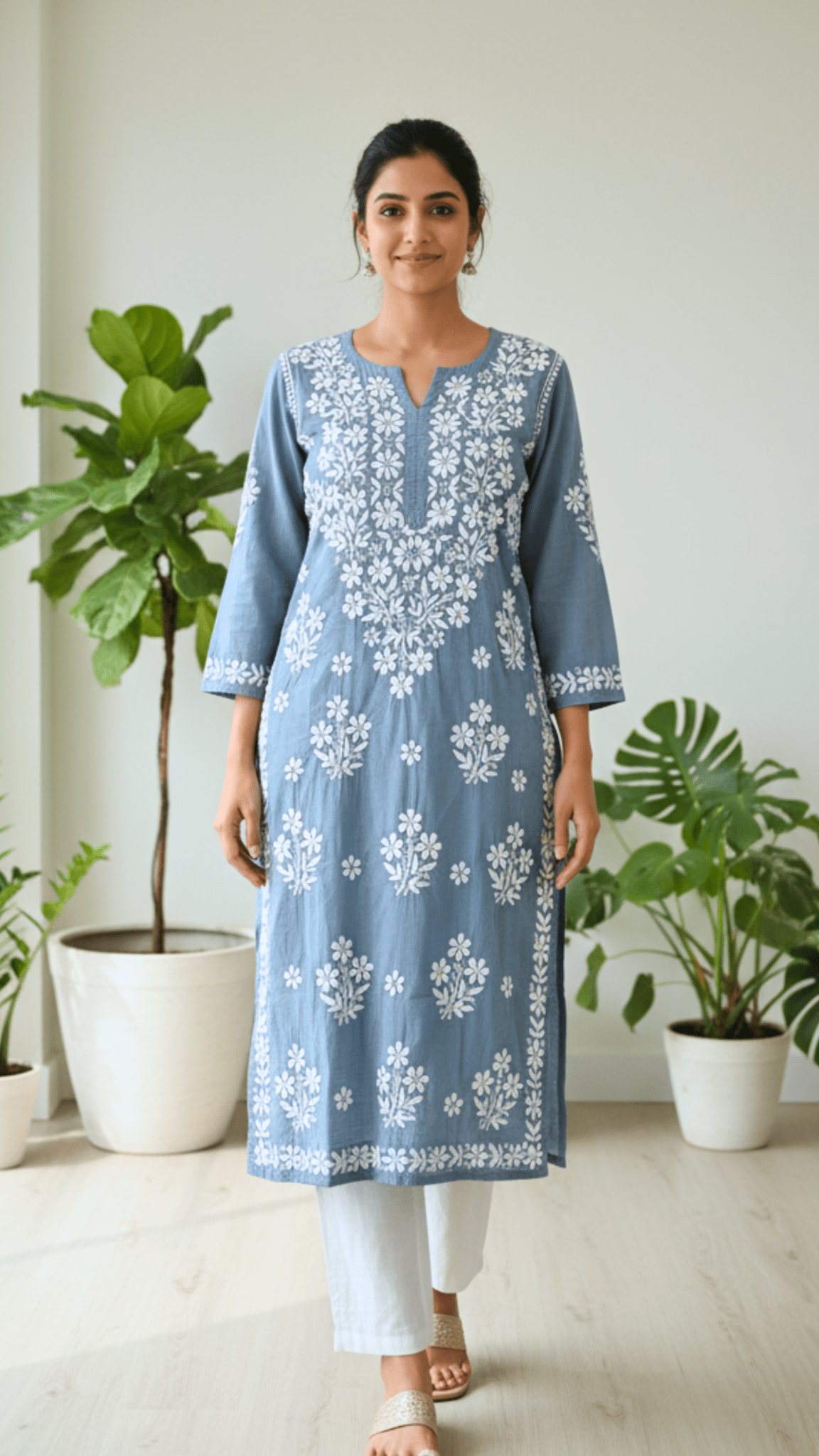 Woman wearing a blue floral chikankari embroidered kurta with white pants in a room with plants.