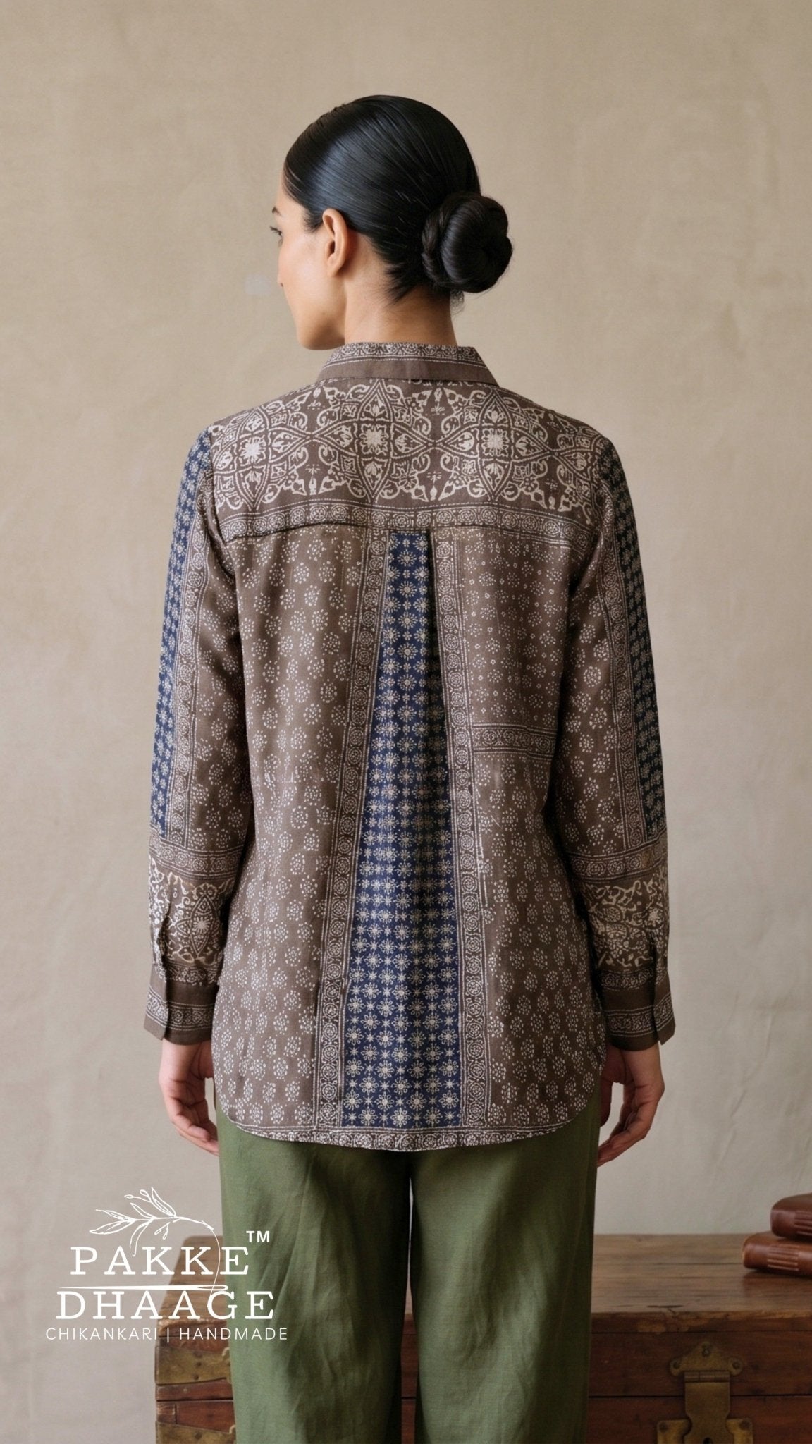 A woman shown from the back wearing a brown and navy patterned long-sleeve shirt with green pants, standing against a neutral beige wall. The brand name 'Pakke Dhaage' is visible as a watermark in the bottom-left corner.