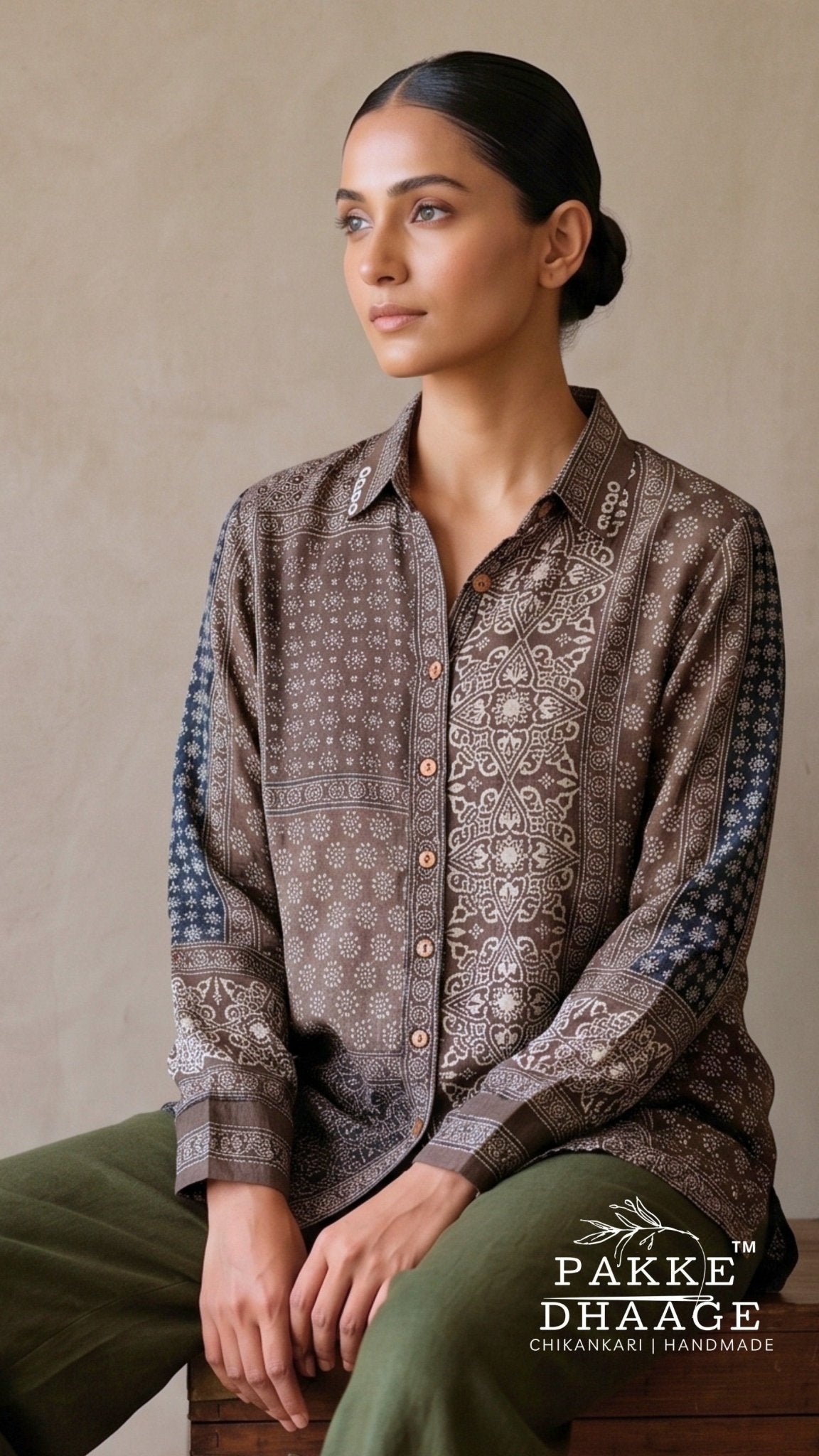 A woman seated against a neutral beige wall wearing a long-sleeve button-front shirt with an allover brown and beige geometric print and blue patterned sleeve panels, paired with olive green pants. Pakke Dhaage