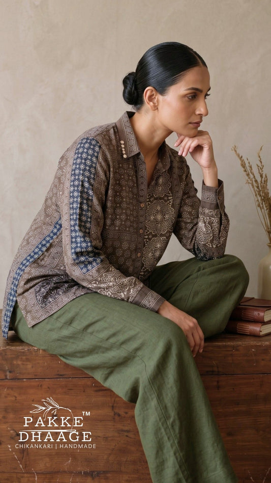 A woman sits on a rustic wooden bench wearing a patterned brown and blue long-sleeve shirt with olive green pants. The brand logo 'PAKKE DHAAGE' is visible in the bottom left corner.