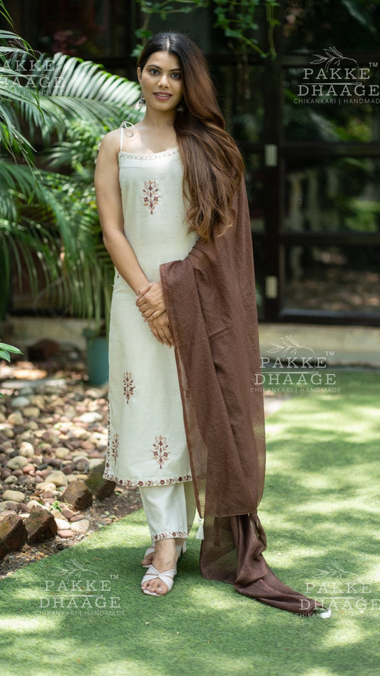 Woman in a white and brown tsuit set standing outdoors with greenery in the background.