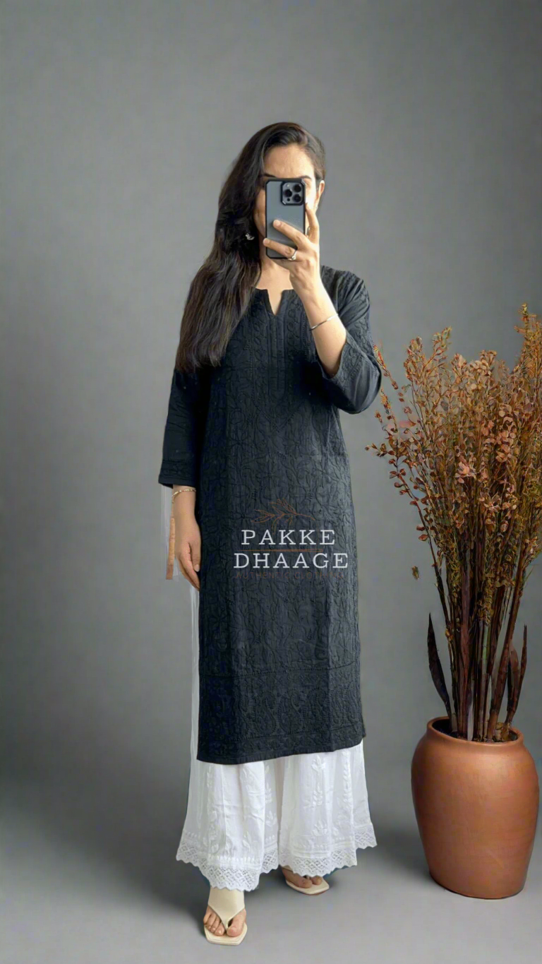 Woman taking a mirror selfie wearing a black chikankari kurta with white pants in a room with plants.