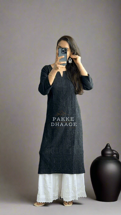 Person wearing a black chikankari kurta with white pants taking a mirror selfie in a room with plants.