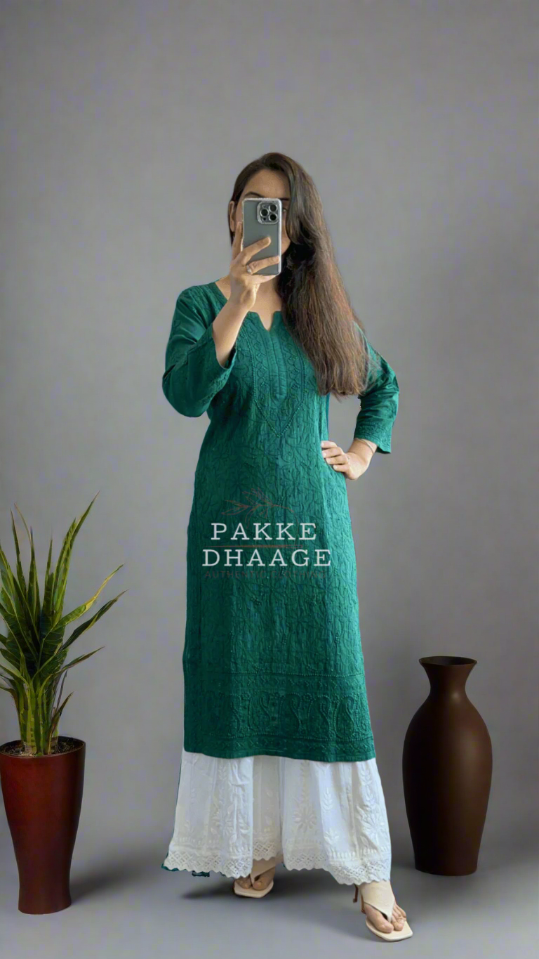Person wearing a green chikankari kurti with white pants standing in a room with plants.