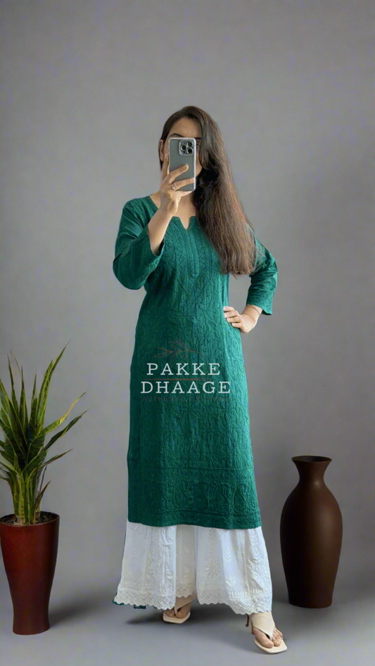 Person wearing a green chikankari kurti with white pants standing in a room with plants.