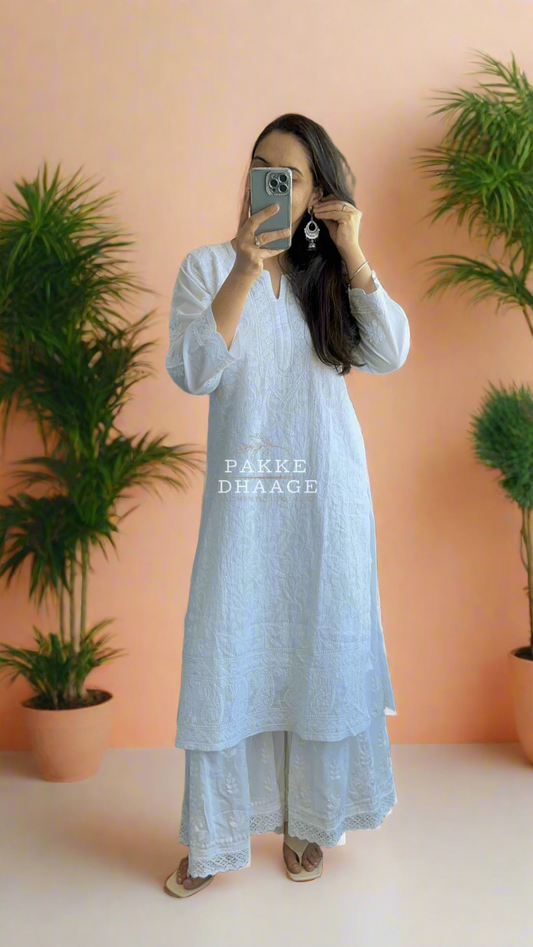 Person wearing a white Chikankari outfit taking a mirror selfie on a balcony with plants.