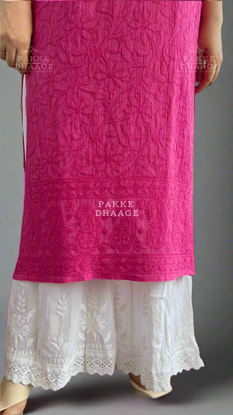 Person wearing a pink embroidered kurta with white pants, standing indoors with plants in the background.