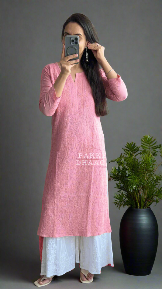 Person wearing a pink chikankari kurta with white pants standing in front of a mirror on a balcony with plants.