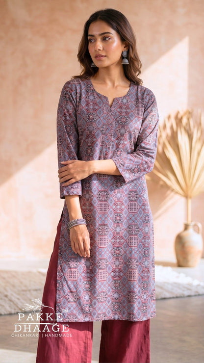 A woman models a traditional Indian outfit from Pakke Dhaage, featuring a lavender-purple printed kurta with red and white geometric patterns, paired with solid maroon pants. The background is a warm, textured beige wall with soft shadows and a decorative dried palm leaf in a vase.
