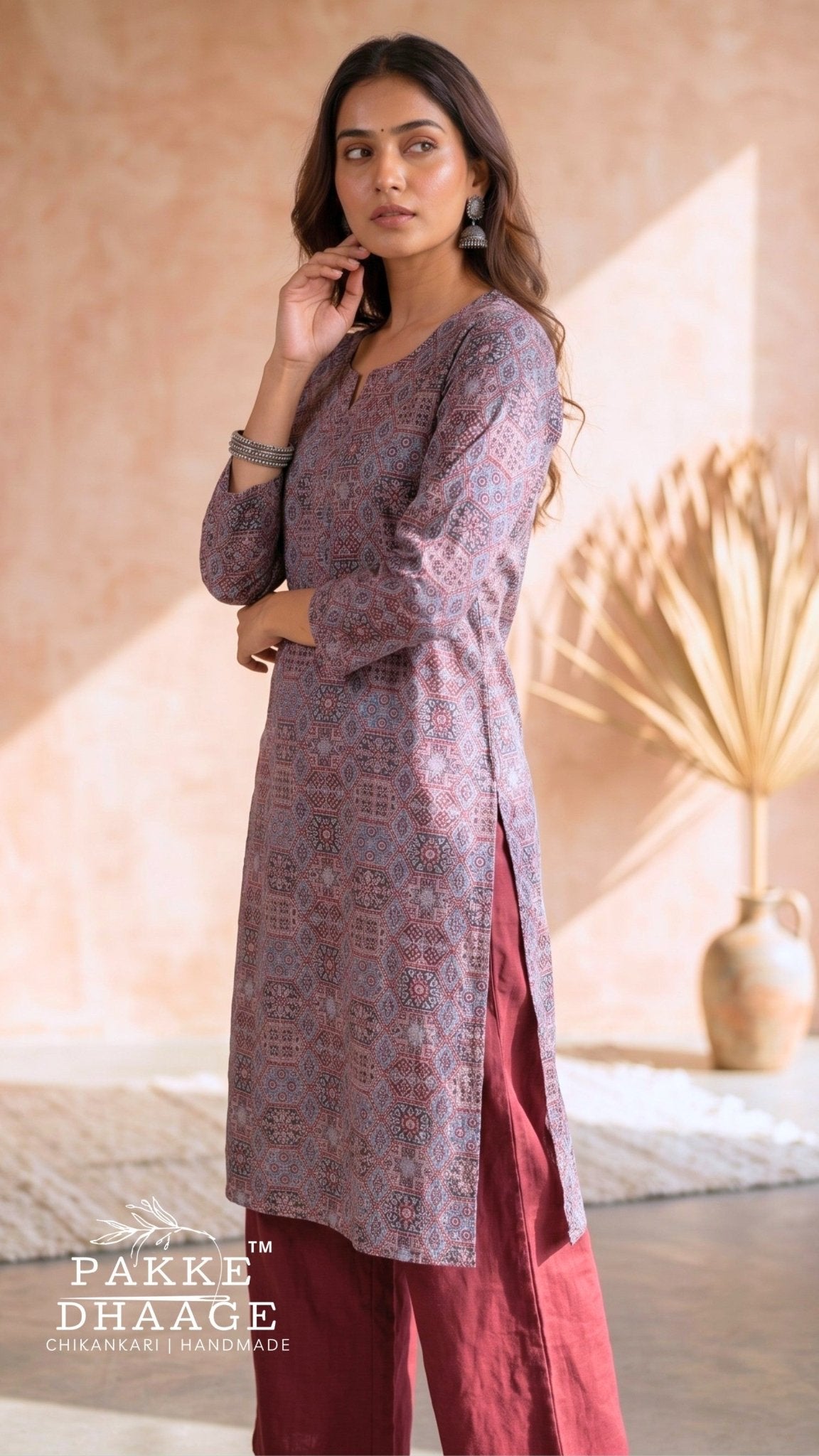 A woman models a three-piece traditional Indian outfit from Pakke Dhaage, featuring a dusty purple printed kurta, a deep red skirt, and matching red trousers, set against a warm beige background with a dried palm leaf decor. | Pakke Dhaage