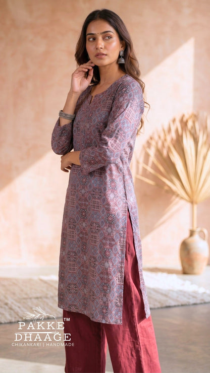A woman models a three-piece traditional Indian outfit from Pakke Dhaage, featuring a dusty purple printed kurta, a deep red skirt, and matching red trousers, set against a warm beige background with a dried palm leaf decor. | Pakke Dhaage