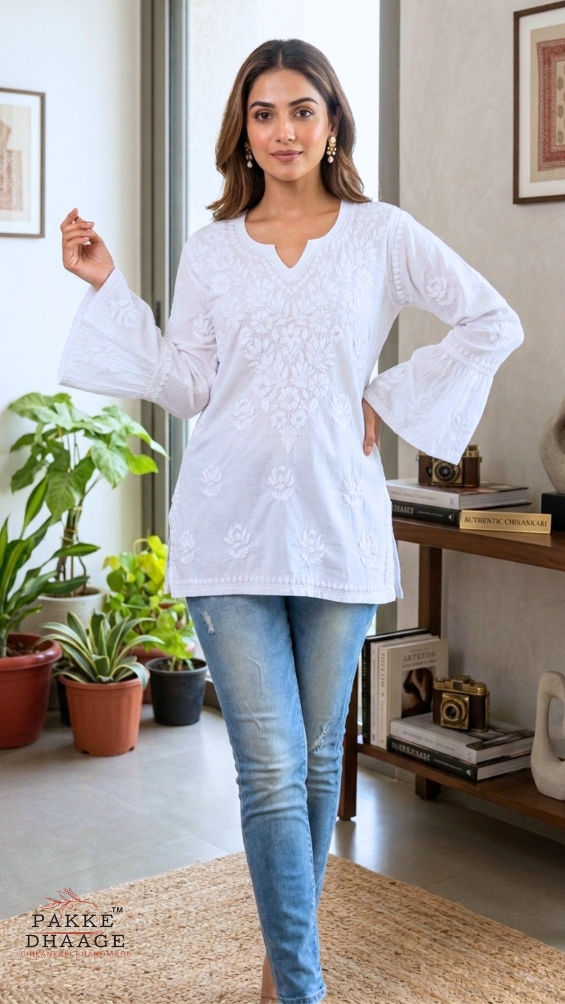 Woman wearing a white chikankari embroidered top and blue jeans in a room with plants and books.