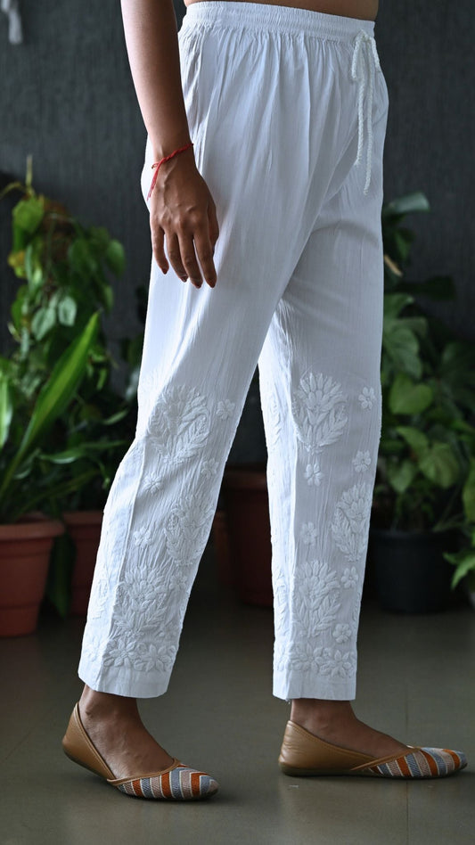 White embroidered cotton trousers with floral detailing on the lower legs, shown on a model wearing brown flat shoes in front of a dark wall with potted plants.