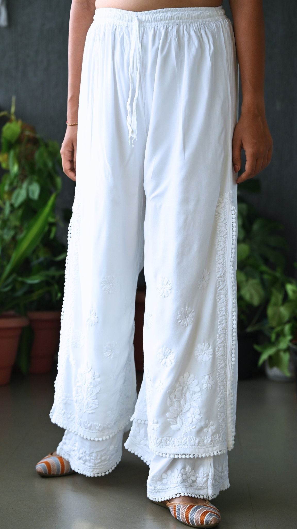 White wide-leg chikankari embroidered pants with a drawstring waist, shown on a model from the waist down in front of a dark gray wall with potted green plants. | Pakke Dhaage