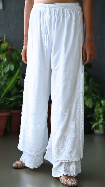 White wide-leg chikankari embroidered pants with a drawstring waist, shown on a model from the waist down in front of a dark gray wall with potted green plants. | Pakke Dhaage