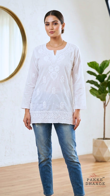 Woman wearing a chikankari white embroidered top and blue jeans standing in a room with a plant and mirror.