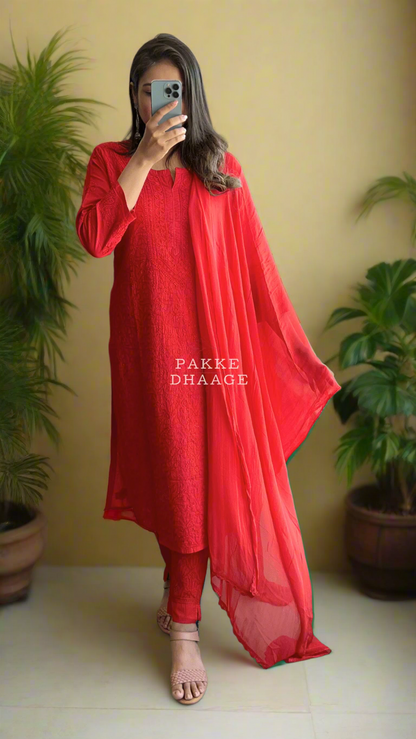 Person wearing a red chikankari kurti set with matching dupatta with a sheer dupatta, standing in front of a mirror on a balcony.