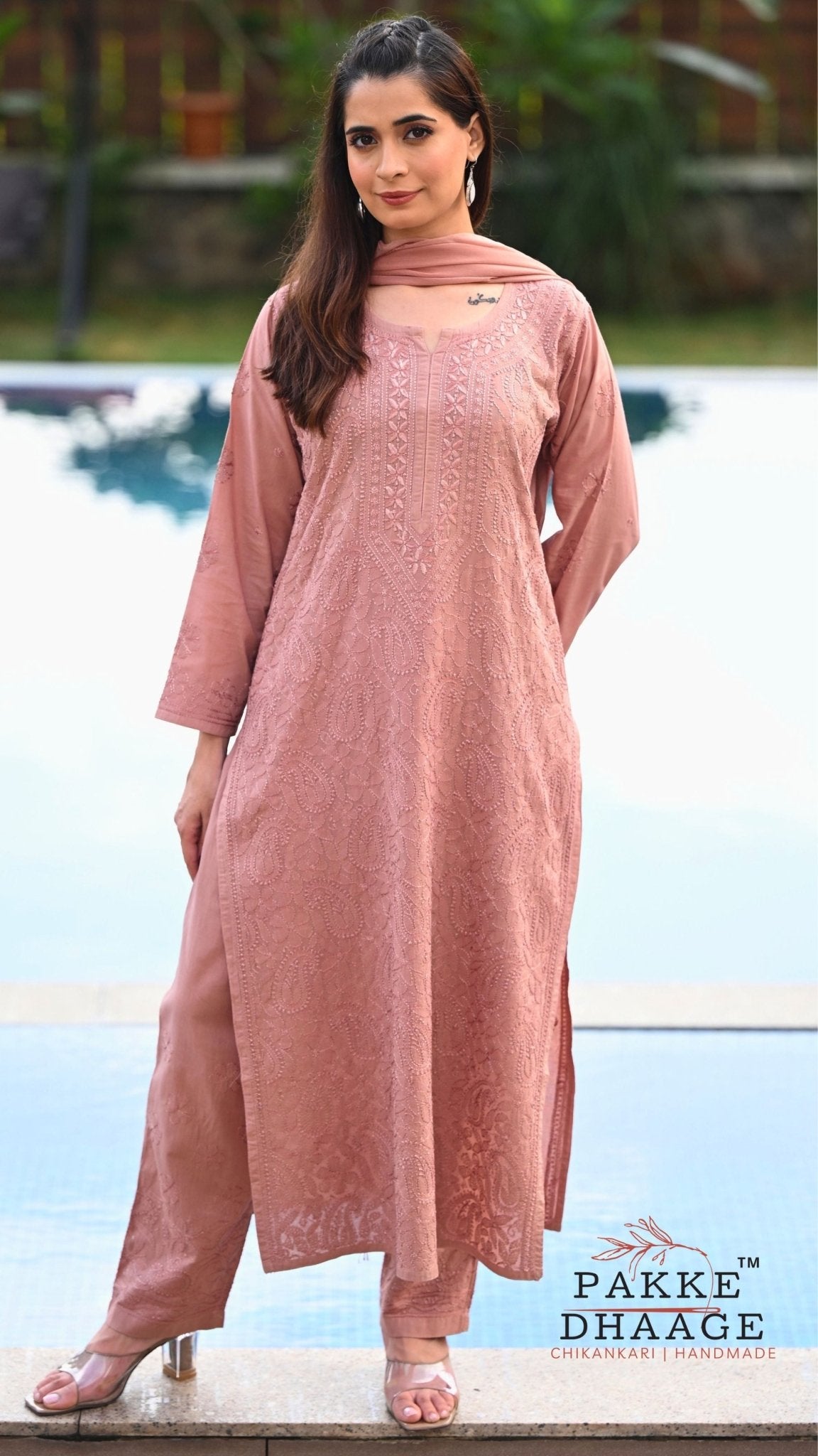 A woman wearing a dusty rose pink embroidered kurta set with matching trousers and a dupatta, standing by a swimming pool. The brand logo 'PAKE DHAAGE' is visible in the bottom right corner.