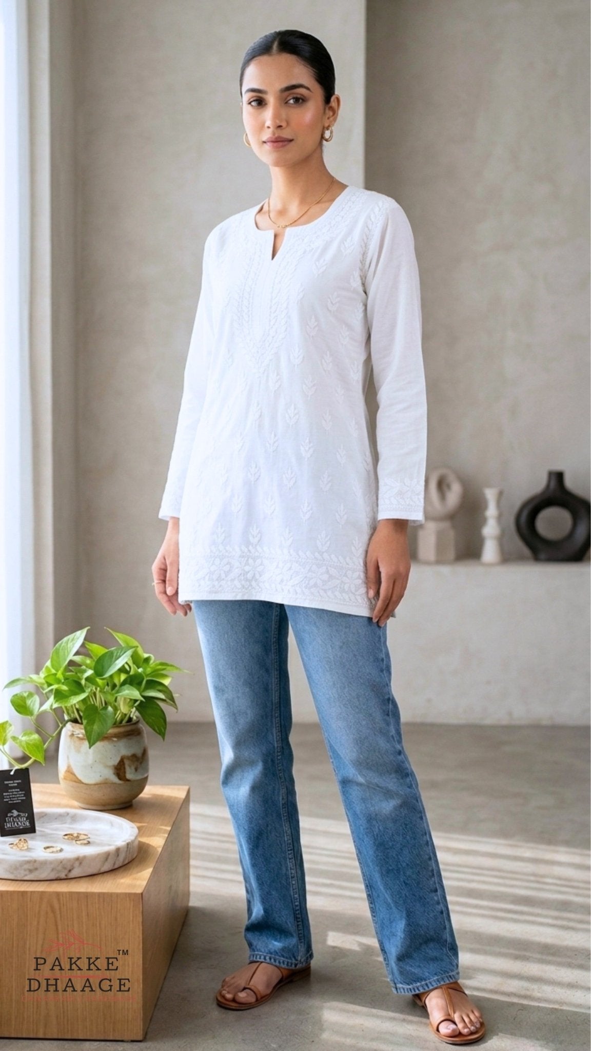 Woman wearing a white chikankari top and blue jeans standing in a room with decor elements.