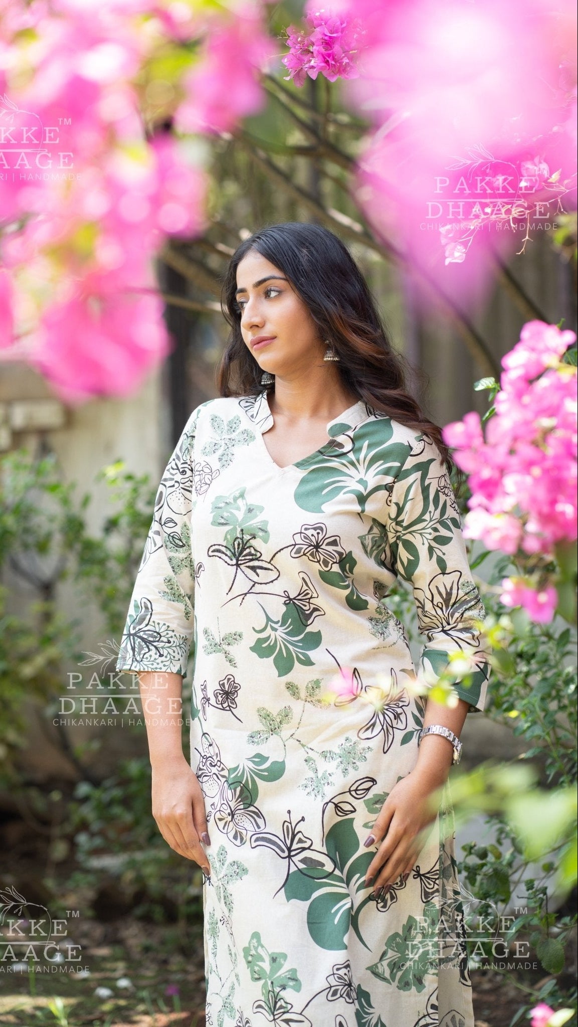 Woman in a floral kurti set standing among pink flowers with 'Pakke Dhaage' branding.