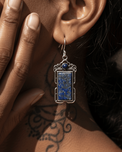 Earring with blue stone and silver frame worn by a person with tattoos on their arm.