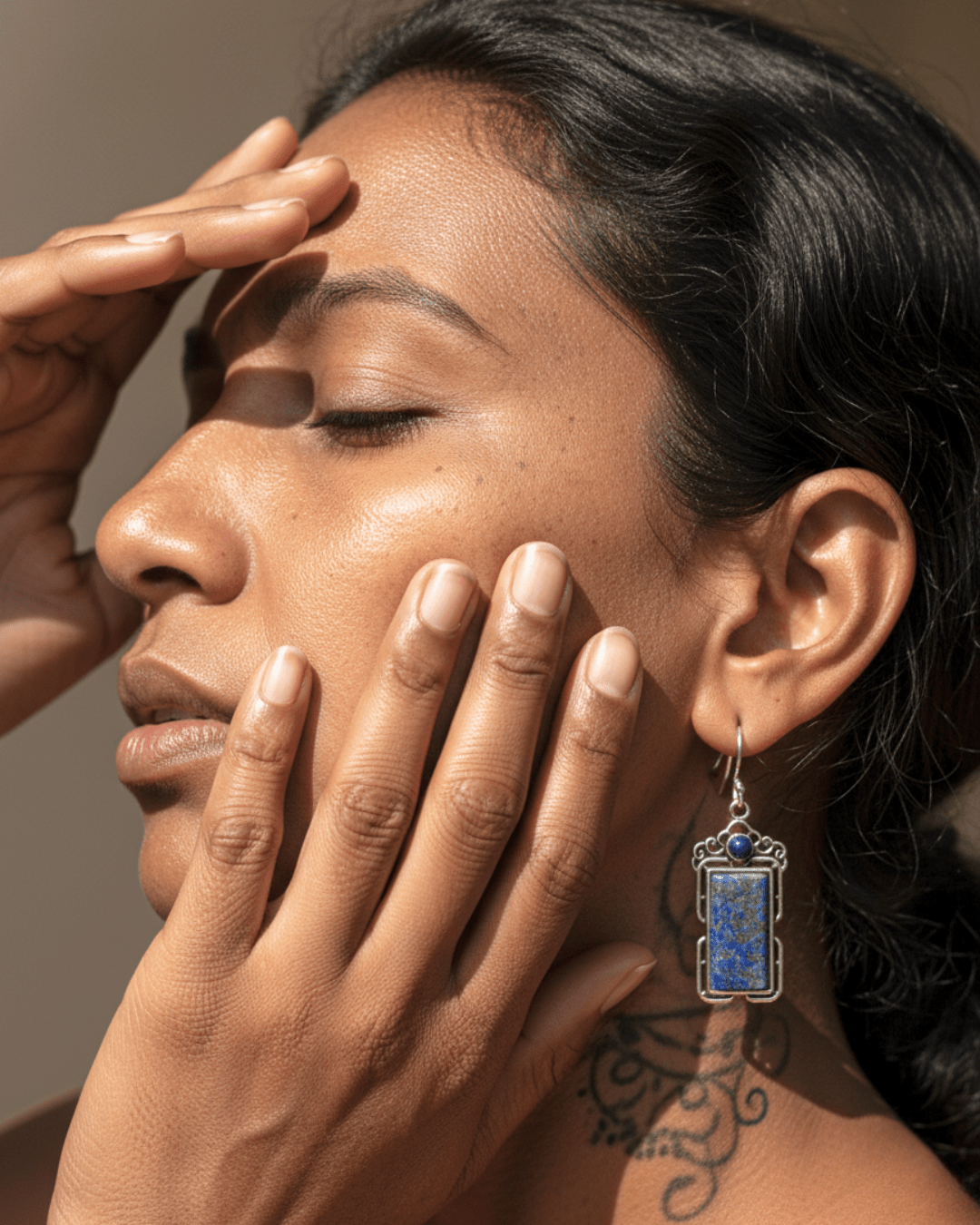 Close-up of a woman's face with one hand on her forehead and the other touching her cheek, wearing an earring.