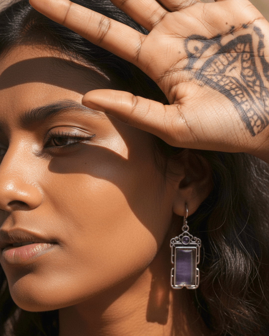 Close-up of a woman's face with a hand touching her forehead, wearing a purple handmade oxidized earring.