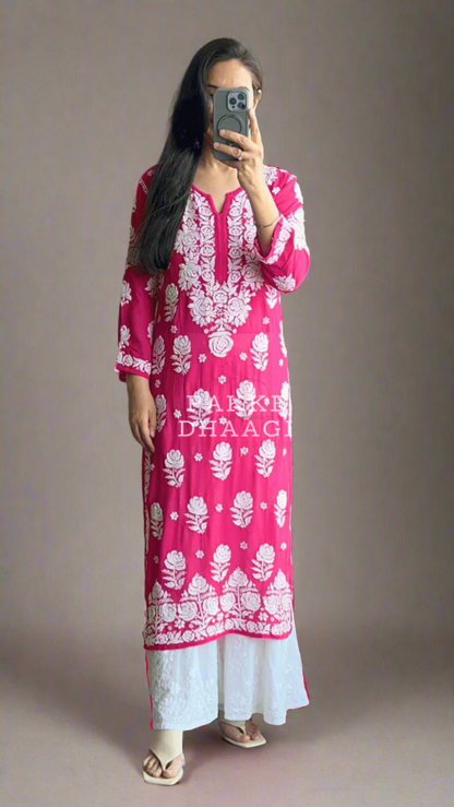 Person wearing a pink and white patterned chikankari kurti standing in front of a mirror with plants around.