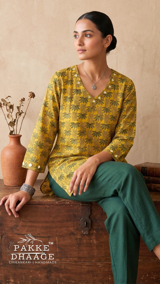 A woman models a yellow and green printed kurta with green trousers, sitting on a rustic wooden chest in a warm, earthy setting. The brand logo 'Pakke Dhaage' is visible in the bottom left corner.