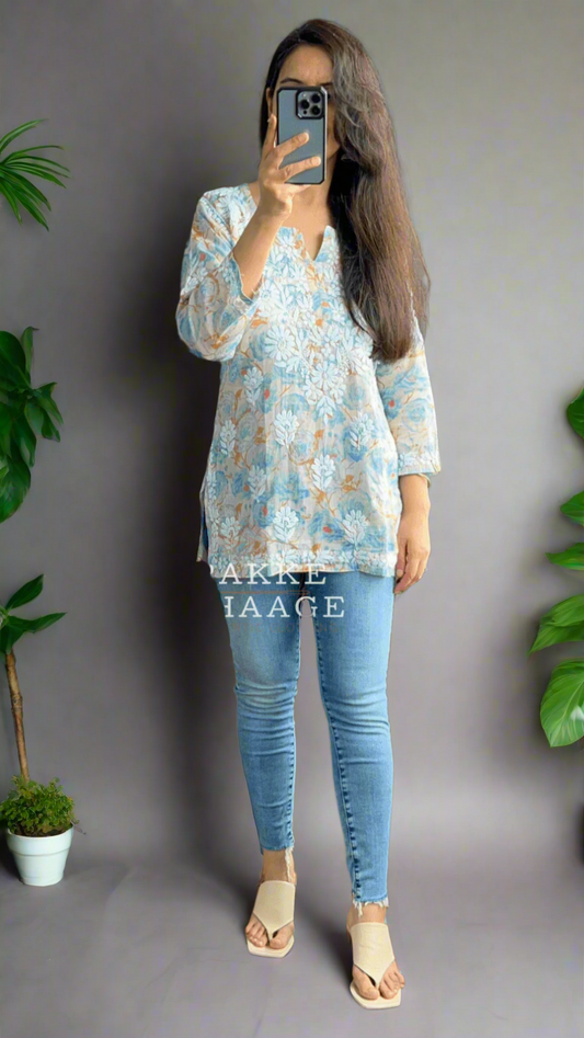 Person taking a mirror selfie wearing a floral chikankari top and blue jeans in a room with plants.