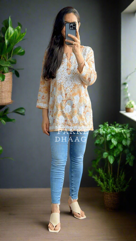 Person taking a mirror selfie wearing a patterned chikankari yellow top and blue jeans in a room with plants.