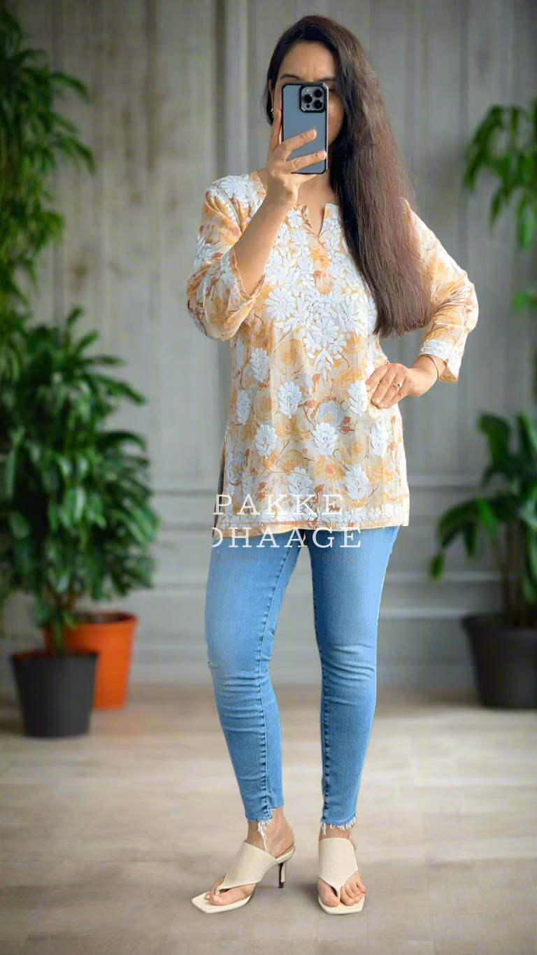 Person taking a mirror selfie wearing a floral patterned printed chikankari top and blue jeans in a room with plants.