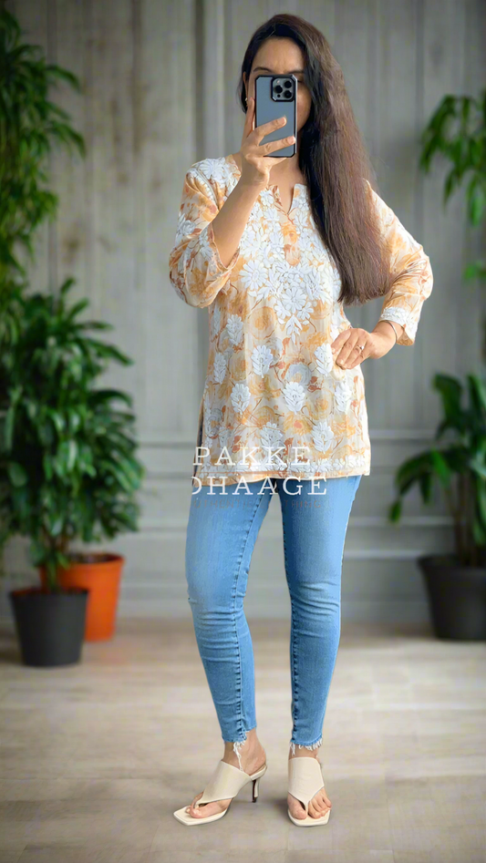 Person taking a mirror selfie wearing a floral patterned printed chikankari top and blue jeans in a room with plants.
