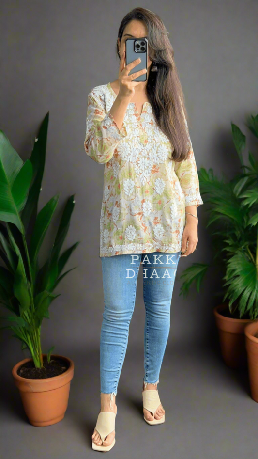 Woman taking a mirror selfie wearing a floral lime colored chikankari top and blue jeans in a room with plants.