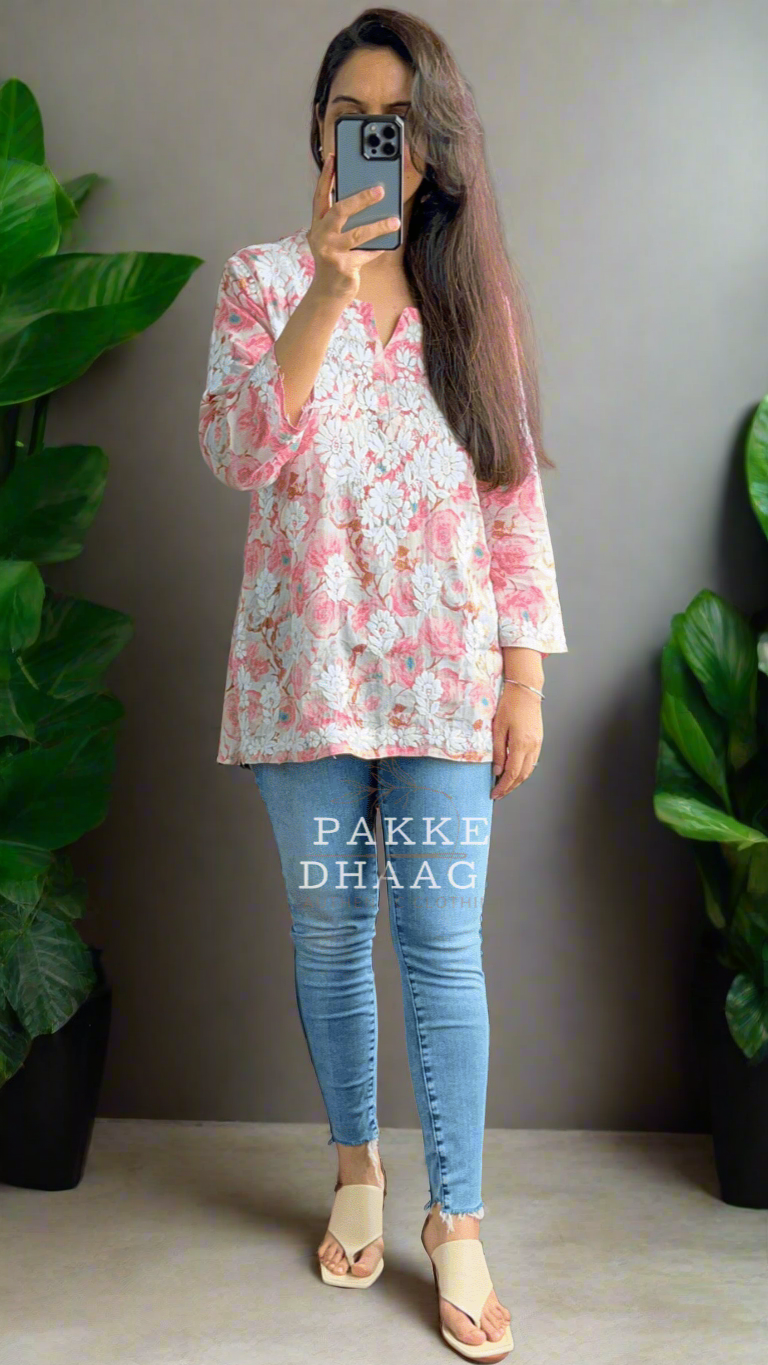Person taking a mirror selfie wearing a floral chikankari printed top and blue jeans in a room with plants.