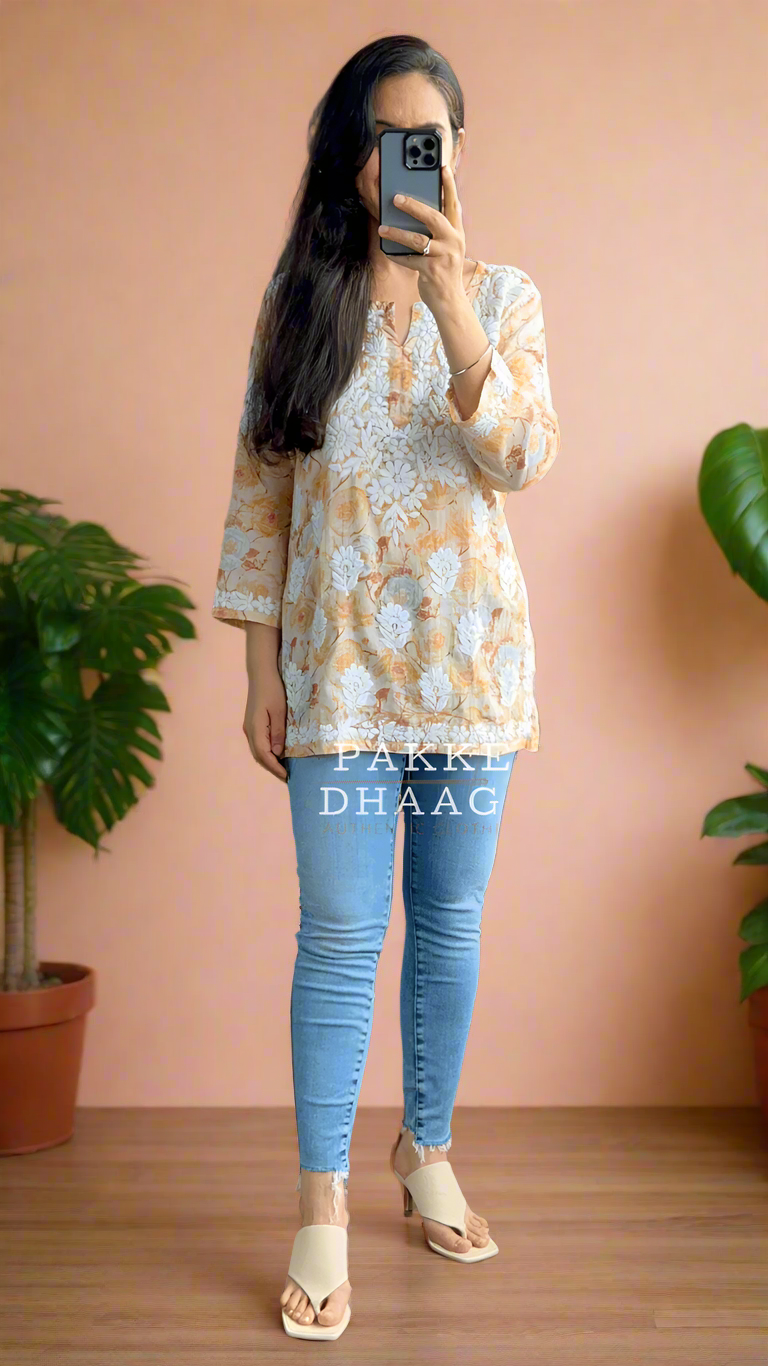 Person taking a mirror selfie wearing a patterned chikankari top and blue jeans in a room with plants.