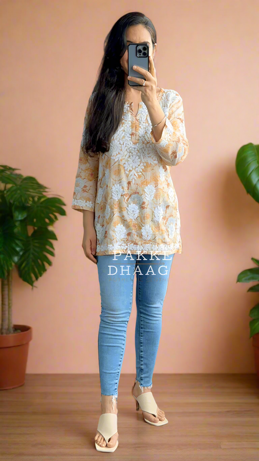 Person taking a mirror selfie wearing a patterned chikankari top and blue jeans in a room with plants.