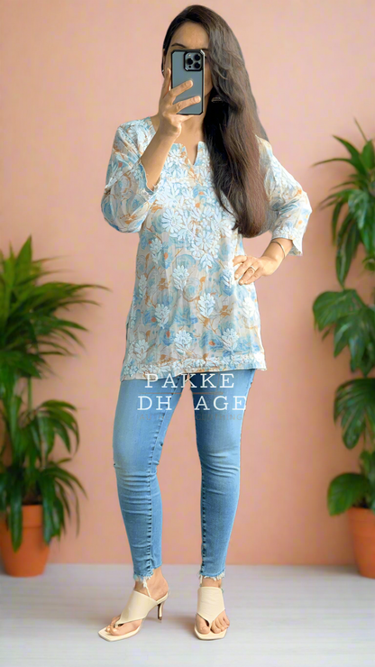 Person taking a mirror selfie wearing a blue patterned chikankari top and light blue jeans in a room with plants.