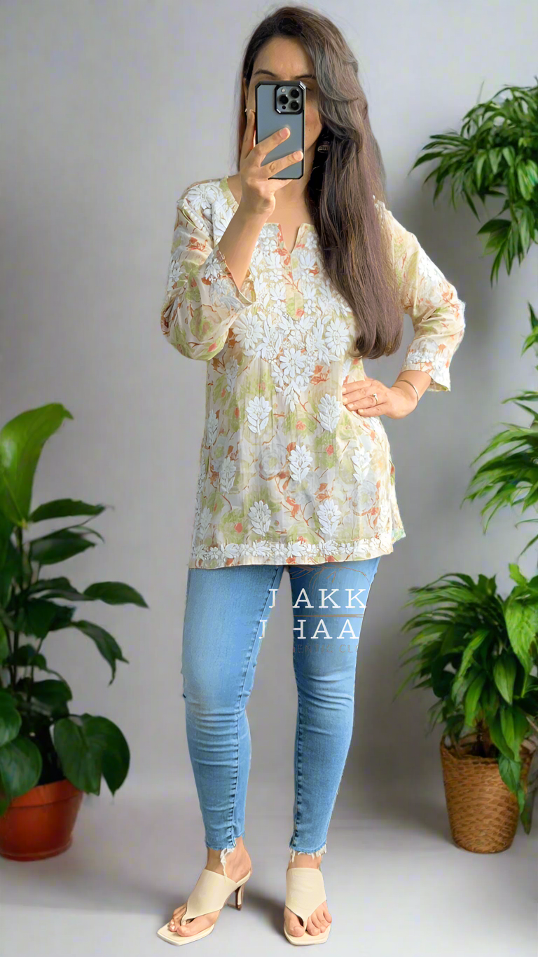 Person taking a mirror selfie wearing a floral chikankari top and blue jeans in a room with plants.